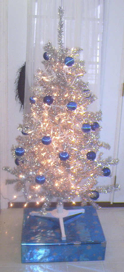 An aluminum Christmas tree with white lights and blue satin ball ornaments, sitting on a blue box.