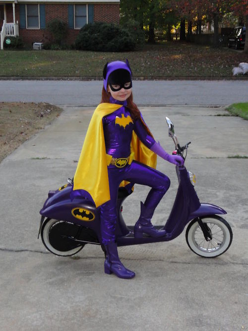 A little girl in a very professionally done Batgirl costume, sitting on a purple scooter with Batman insignia.