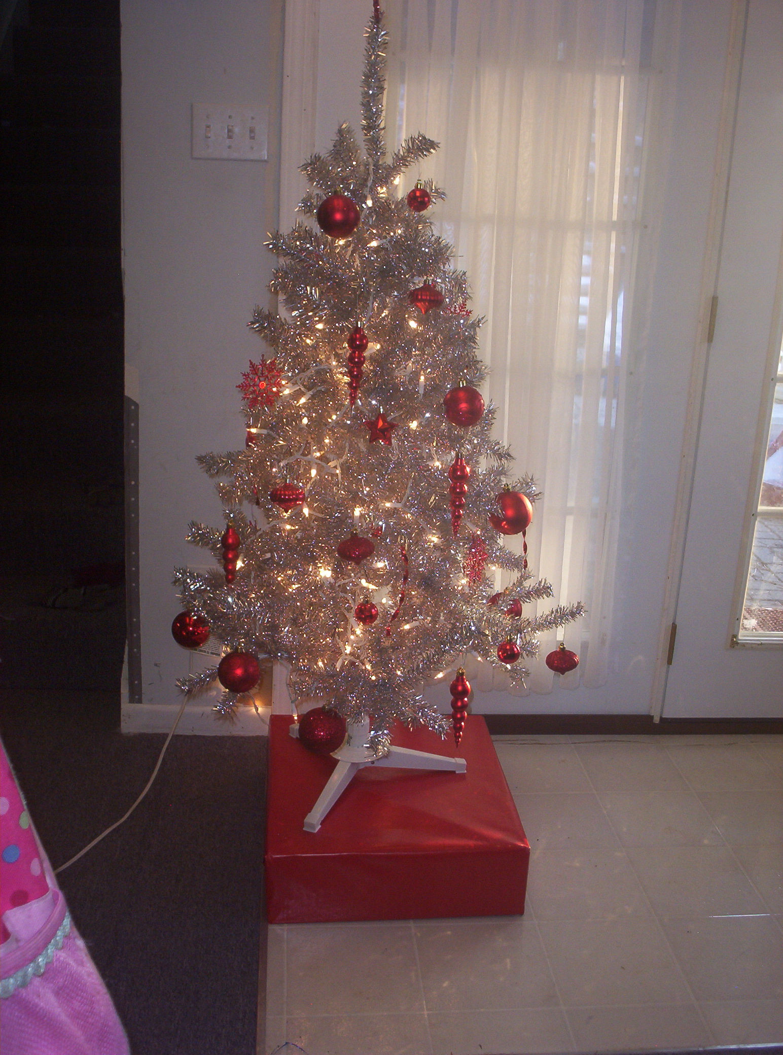 A small aluminum Christmas tree with white lights and red ornaments, standing on a red box.