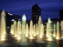 Nighttime. A flat expanse of concrete with many equally spaced jets of water, with a skyscraper in the background.