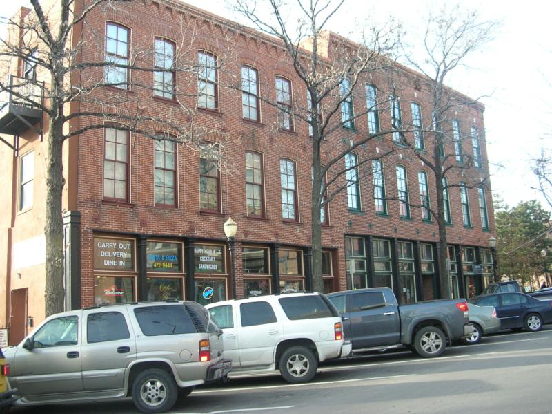 A 19th-century 3-story brick building with shops on the bottom, with cars parked in front.