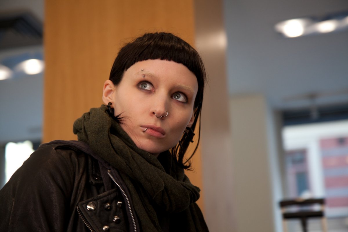 A still shot of Rooney Mara in THE GIRL WITH THE DRAGON TATTOO. She has a very short black haircut with a straight line of bangs, and piercings on her face. She's wearing a black scarf and jacket.