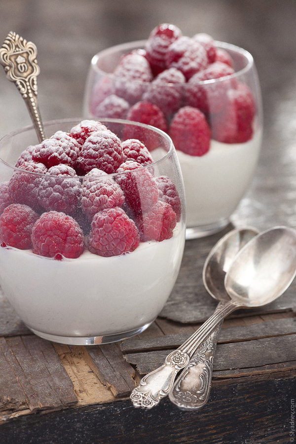 An image of cups of yogurt topped with sugared or iced raspberries, on a weathered table and mismatched silver spoons of vintage patterns.
