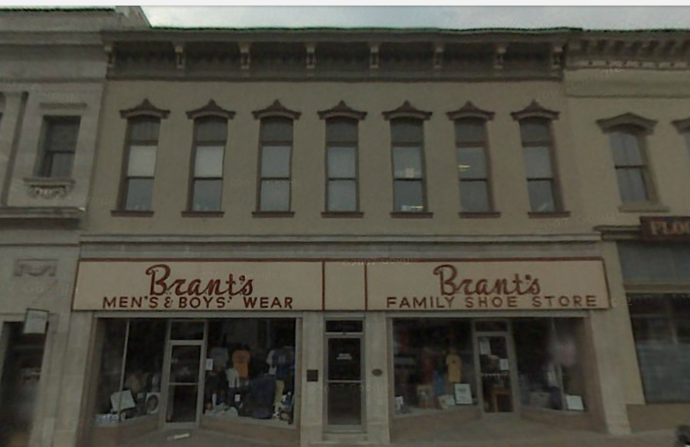 A 19th-century 2-story cream stucco building on an American town square surrounding a courthouse that says Brant’s Men’s & Boys’ Wear.
