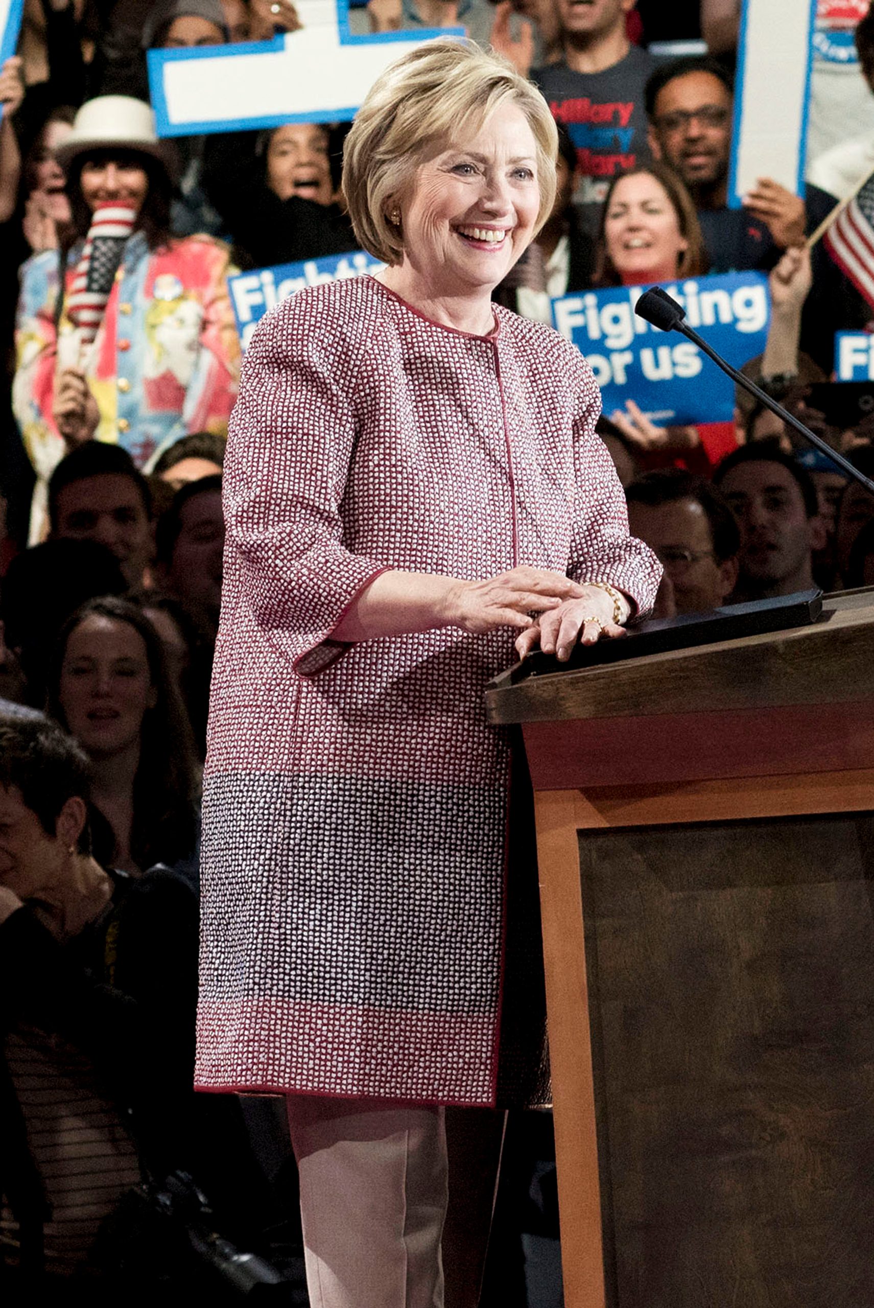 An image showing Hillary Clinton behind a podium with a mid-thigh-length red, black, and white jacket that looks crocheted.