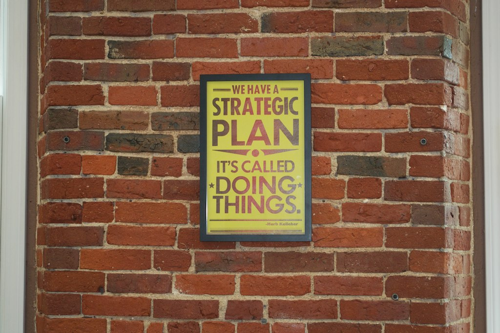 An image of a poster hanging on a brick wall that says “We have a strategic plan. It's called DOING THINGS.”