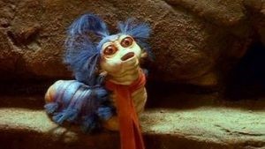 An image of a tiny muppet worm with tiny blue feathers sprouting out of his head and a tiny red scarf.