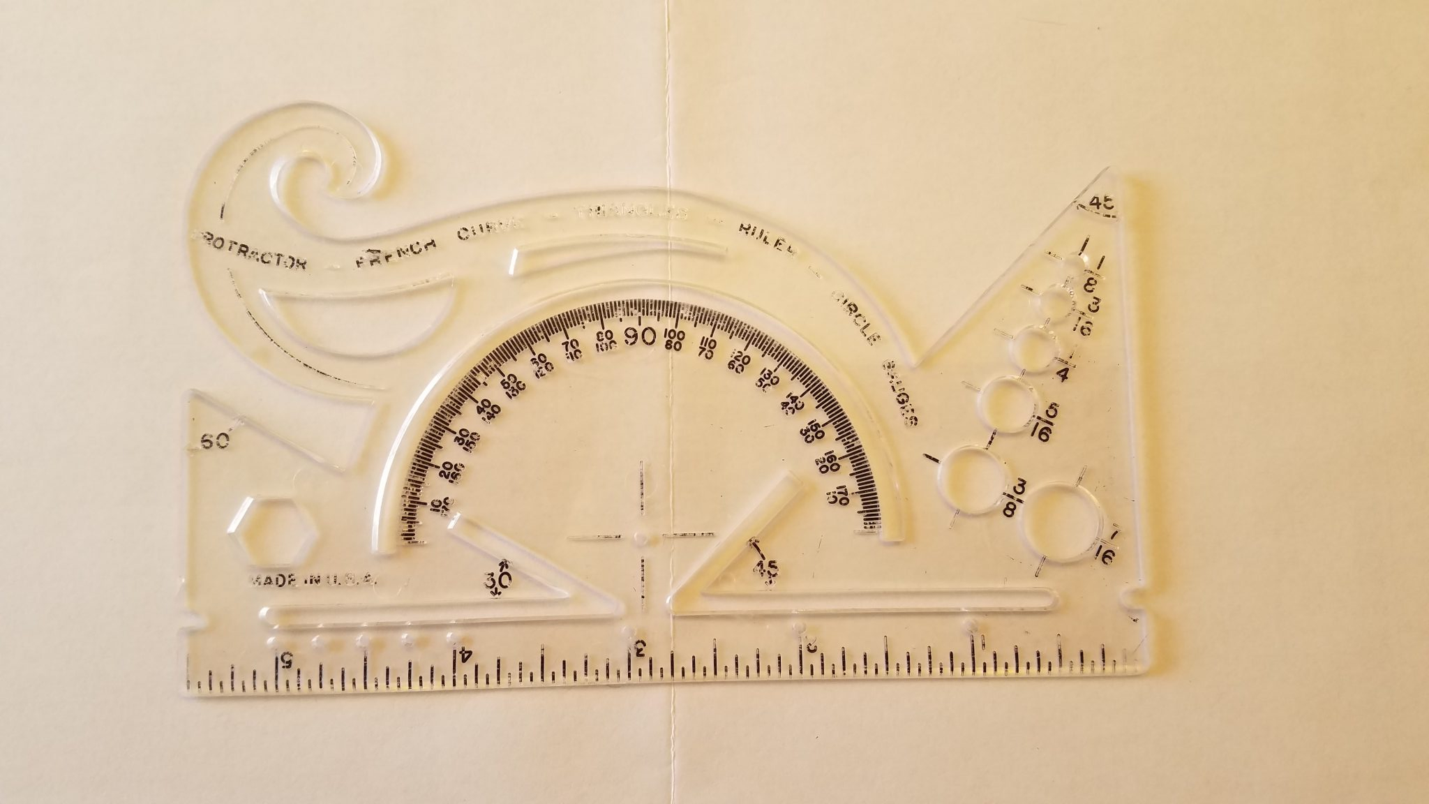An image of a small old-school (1980s) plastic combination protractor, ruler, French curve, circle gauges, and triangle and hexagon and line templates,