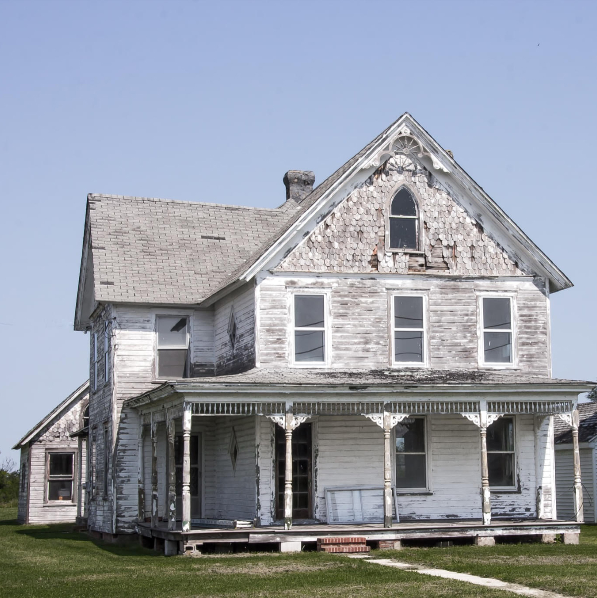 A filthy, dilapidated white two-story 19th-century gothic revival farmhouse with two one-story outbuildings.